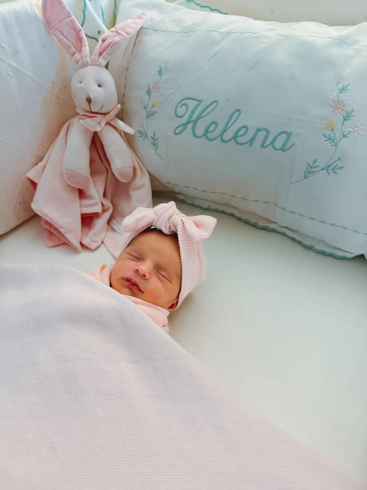 Baby Helena asleep in a pink bow headband, beside a pillow embroidered with her name